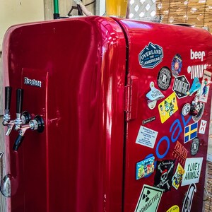 May include: A red vintage refrigerator with a beer tap and various stickers, including "Overland the Americas", "Bonfire", "Penguin Coffee", and "Live 1 Animals". A glass of beer sits on top of the refrigerator.