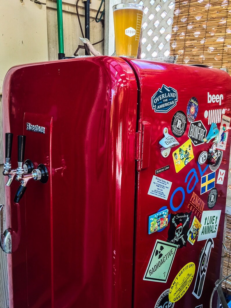 May include: A red vintage refrigerator with a beer tap and various stickers, including "Overland the Americas", "Bonfire", "Penguin Coffee", and "Live 1 Animals". A glass of beer sits on top of the refrigerator.