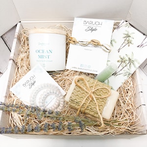 Könnte beinhalten: Eine Geschenkbox mit einer weißen Kerze mit der Aufschrift "Ocean Mist", einem goldenen Muschelarmband, Seife, Haargummis, Lippenbalsam und Lavendel. Die Box ist mit Stroh gefüllt und enthält Karten mit dem "Baruch Style"-Logo.