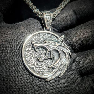 Könnte beinhalten: Eine silberne Halskette mit Anhänger, die ein detailliertes Wolfskopf-Design in einem kreisförmigen Rahmen zeigt. Der Anhänger hängt an einer geflochtenen Kette. Der Wolfskopf ist stilisiert und strukturiert, mit offenem Maul und Zähnen.