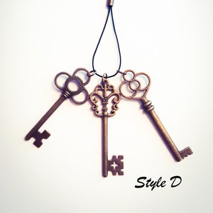 May include: A set of three bronze-toned metal keys attached to a black cord with a lobster clasp. The keys feature intricate, ornate bow designs and distinct bit patterns at the ends. The text "Style D" is printed in black in the bottom right corner.