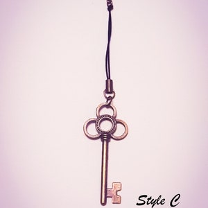 May include: A bronze-colored metal key charm featuring a clover-shaped bow with a central circular cutout, attached to a black cord with a lobster clasp. The text "Style C Cord" is printed in black in the lower right corner of the image.
