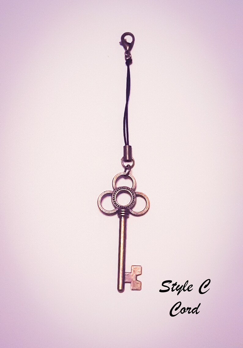 May include: A bronze-colored metal key charm featuring a clover-shaped bow with a central circular cutout, attached to a black cord with a lobster clasp. The text "Style C Cord" is printed in black in the lower right corner of the image.
