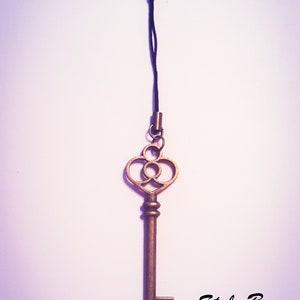 May include: A bronze-colored metal key charm attached to a thin black cord with a lobster claw clasp. The key features an ornate bow with circular cutouts and a straight shaft. The text "Style B Cord" is printed in black in the lower right corner.