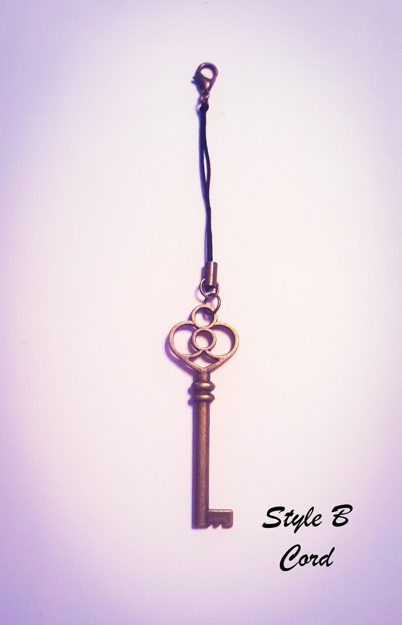 May include: A bronze-colored metal key charm attached to a thin black cord with a lobster claw clasp. The key features an ornate bow with circular cutouts and a straight shaft. The text "Style B Cord" is printed in black in the lower right corner.