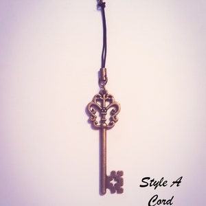 May include: A bronze-colored metal key charm featuring an ornate, symmetrical bow and a square-cut bit. The key is attached to a thin black cord with a small lobster clasp at the top. The text "Style A Cord" is printed in black to the right of the key.