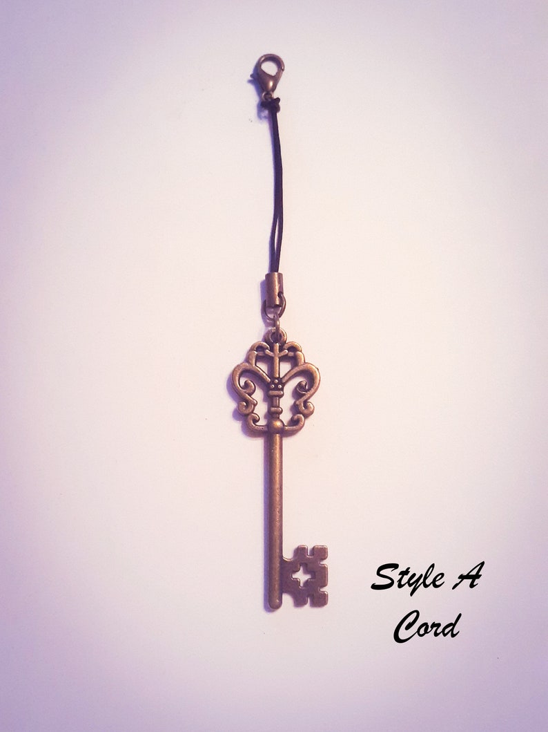 May include: A bronze-colored metal key charm featuring an ornate, symmetrical bow and a square-cut bit. The key is attached to a thin black cord with a small lobster clasp at the top. The text "Style A Cord" is printed in black to the right of the key.