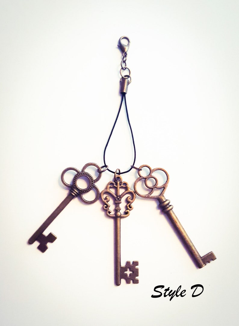 May include: A set of three bronze-toned metal keys attached to a black cord with a lobster clasp. The keys feature intricate, ornate bow designs and distinct bit patterns at the ends. The text "Style D" is printed in black in the bottom right corner.