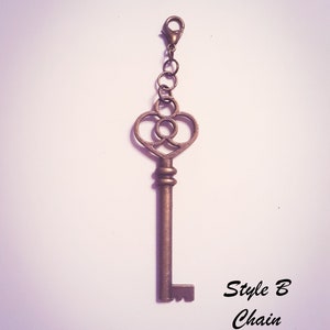May include: A bronze-toned metal key charm featuring a heart-shaped bow with a circular cutout center. The key is attached to a short chain with a lobster claw clasp. The text "Style B Chain" is printed in black in the lower right corner.