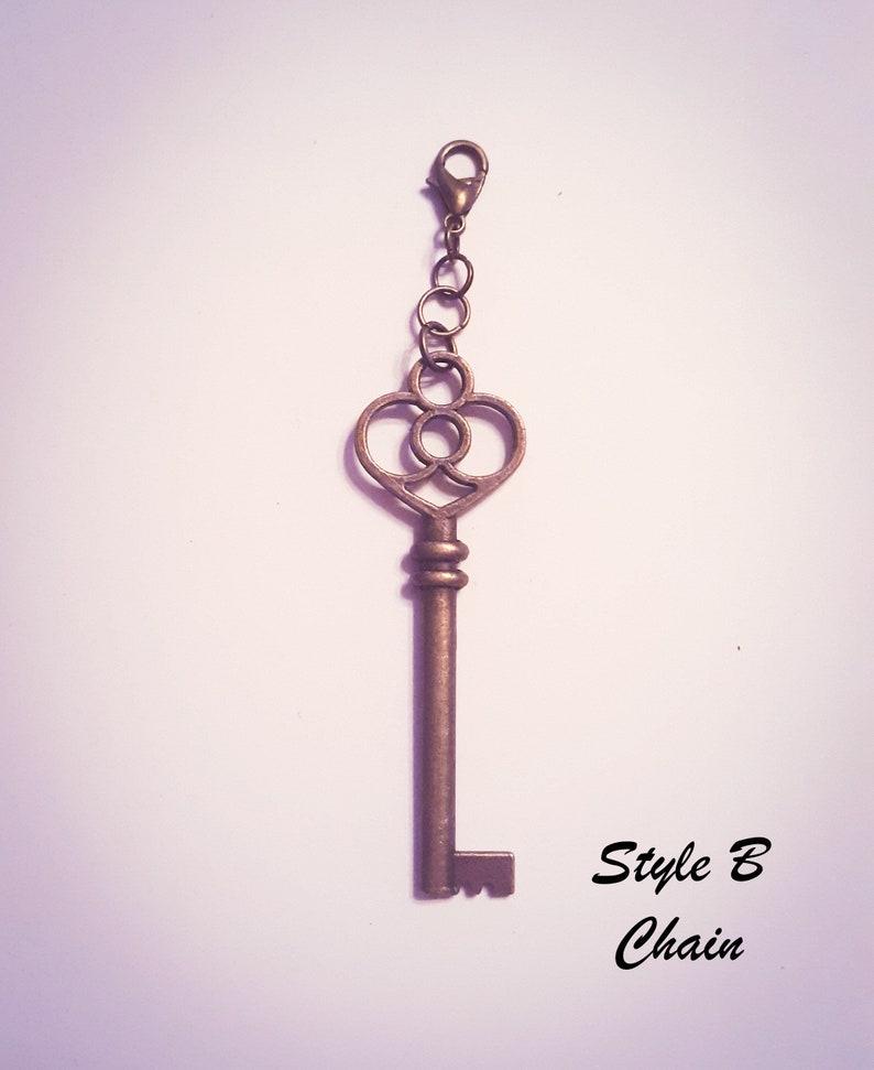 May include: A bronze-toned metal key charm featuring a heart-shaped bow with a circular cutout center. The key is attached to a short chain with a lobster claw clasp. The text "Style B Chain" is printed in black in the lower right corner.
