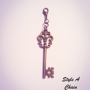 May include: A bronze-colored metal key charm featuring an ornate, openwork bow and a bit with a cross-shaped cutout. The key is attached to a small lobster clasp by two jump rings. The text "Style A Chain" is printed in black in the lower right corner.