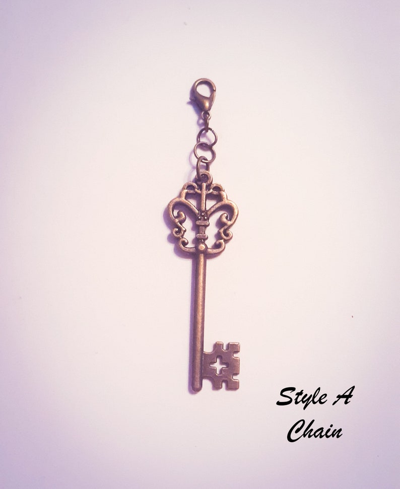 May include: A bronze-colored metal key charm featuring an ornate, openwork bow and a bit with a cross-shaped cutout. The key is attached to a small lobster clasp by two jump rings. The text "Style A Chain" is printed in black in the lower right corner.