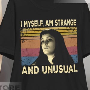 May include: Black t-shirt with the text "I MYSELF, AM STRANGE AND UNUSUAL" and a portrait of a woman. The text is in white, with a retro sunset design in the background. The shirt is hanging on a wooden hanger.