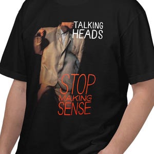 May include: A black short-sleeved t-shirt featuring a graphic print of a person in a large beige suit. The text "TALKING HEADS" appears in white, while the words "STOP MAKING SENSE" are printed in orange below the figure on the front of the shirt.