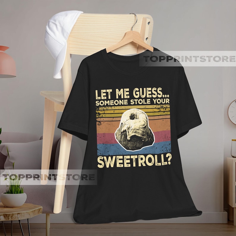 May include: Black t-shirt with a graphic of a sweetroll and text that reads "LET ME GUESS... SOMEONE STOLE YOUR SWEETROLL?" The design features a retro color scheme of orange, yellow, blue, and red stripes.