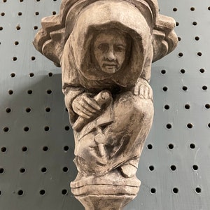 May include: A carved stone sculpture of a hooded figure, possibly a monk or saint, in a seated position. The sculpture is a light gray color and features detailed facial features and clothing. The figure holds an object in its hands. The sculpture has a decorative base.