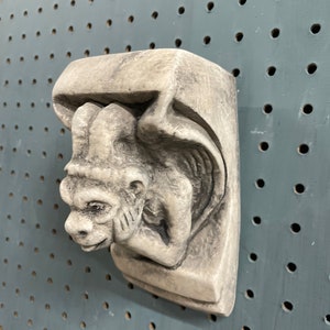 May include: A gray, stone-like gargoyle wall sculpture. The sculpture features a detailed face with pointed ears, a small mouth, and a wing. The gargoyle is mounted on a rectangular base.