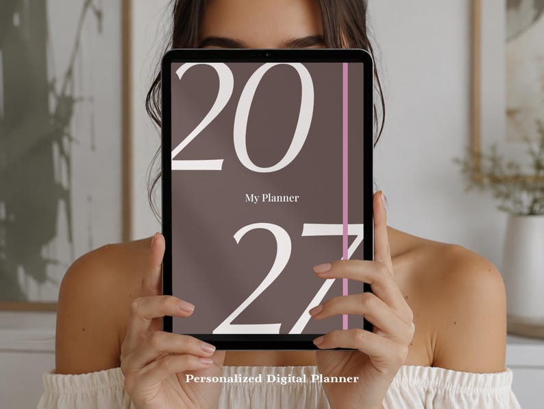 May include: A digital tablet displaying a brown screen with the large white text 2027, the words My Planner in smaller white font, and a thin vertical pink line. The screen also features the text Personalized Digital Planner at the bottom.