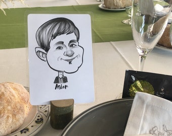Op de afbeelding: Een zwart-witte karikatuur van een persoon met de naam Asier eronder. De karikatuur staat op een witte kaart en staat op een houten standaard. De kaart ligt op een tafel met een bord brood, een vork en een servet.