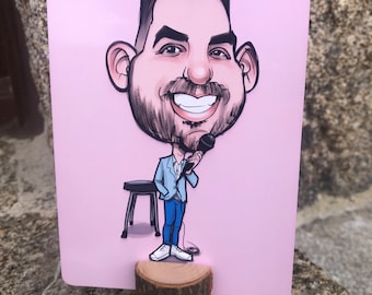Op de afbeelding: Een cartoon-karikatur van een persoon met een microfoon, die een blauwe blazer en jeans draagt, staand op een kleine kruk. De afbeelding is afgedrukt op een roze kaart en gemonteerd op een houten basis.