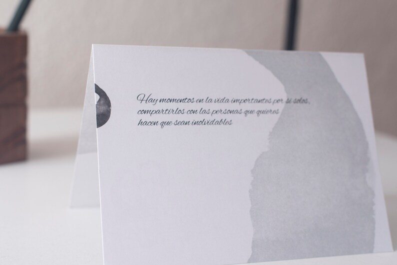 Op de afbeelding: Een witte kaart met een grijze aquarel achtergrond en een citaat in het Spaans dat luidt: "Hay momentos en la vida importantes por s&iacute; solos, compartirlos con las personas que quieres hacen que sean inolvidables."