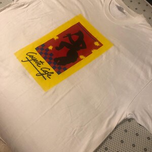 Puede incluir: Una camiseta blanca de manga corta con un estampado gráfico rectangular. El diseño incluye un borde amarillo, un centro rojo con la silueta negra de un flautista, un patrón de cuadros azul y rojo y el texto Coyote Cafe en color negro.
