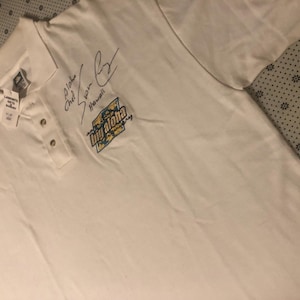 Puede incluir: Un polo blanco de manga corta con cuello y dos botones. En la zona del pecho presenta una firma y un parche gráfico con el texto Big Aloha Hawaii. Una pequeña etiqueta blanca con texto impreso está sujeta en la zona del cuello.