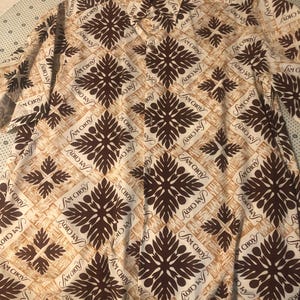 Puede incluir: Una camisa de manga corta con botones que presenta un patrón repetitivo de motivos florales estilizados en marrón oscuro y el texto Sam Choy impreso en letra cursiva sobre un fondo color canela claro con una sutil textura de veta de madera.