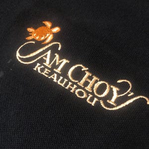 May include: A black polo shirt featuring an embroidered design in gold thread. The embroidery includes a small sea turtle icon above the text SAM CHOY and KEAUHOU, with a decorative flourish extending from the end of the word CHOY.