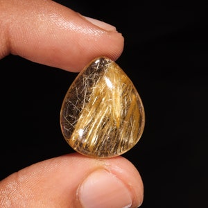 May include: A pear-shaped golden rutilated quartz gemstone featuring a clear, transparent body filled with numerous fine, needle-like golden rutile inclusions that create a distinct, shimmering, and reflective pattern throughout the stone.