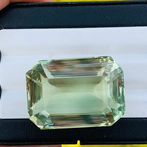 May include: A large, light green gemstone with a rectangular emerald cut. The gemstone is displayed in a black velvet box with a white interior.