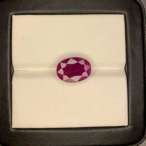 Könnte beinhalten: Ein ovaler Edelstein in tiefem Pink mit facettiertem Schliff, in einer weißen Samt-Präsentationsbox.