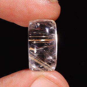 Pode incluir: Um cabochão de quartzo transparente com inclusões de rutilo dourado. O cabochão tem forma retangular e uma superfície lisa e polida.