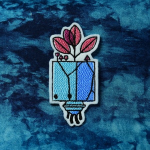 May include: A blue and pink embroidered patch of a plant in a pot with a geometric design. The patch is on a blue and white tie-dye fabric.