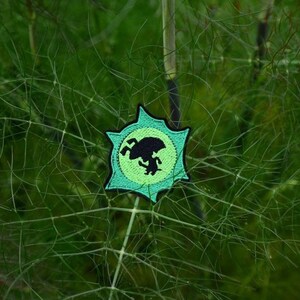 May include: A circular patch featuring a black silhouette of a creature on a light green background, with a darker green border. The patch is set against a backdrop of green foliage.
