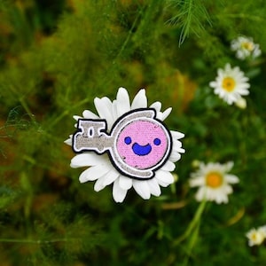 Könnte beinhalten: Ein Aufnäher mit einem lächelnden rosa Charakter mit blauem Mund und Augen, in einem grauen Umriss. Der Aufnäher befindet sich auf einer weißen Gänseblümchen, umgeben von grünem Laub und anderen Gänseblümchen.