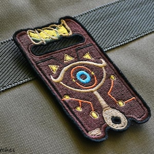 Könnte beinhalten: Ein brauner gestickter Aufnäher mit einem stilisierten Augenmotiv, umgeben von einem Netzwerk aus Linien und Punkten. Das Auge ist blau und orange, mit einem gelben und braunen Rand. Der Aufnäher befindet sich auf einem grauen Stoffhintergrund.