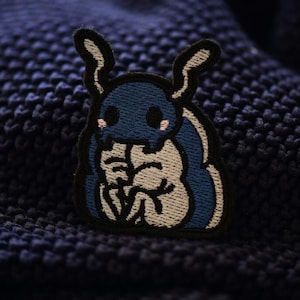 May include: Blue and white embroidered patch of a cartoon creature with large eyes and antennae. The creature is sitting with its arms crossed.