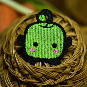 May include: An embroidered patch featuring a cartoon character with a leaf on its head. The character is green with black eyes and pink cheeks. The patch is sitting in a woven basket.