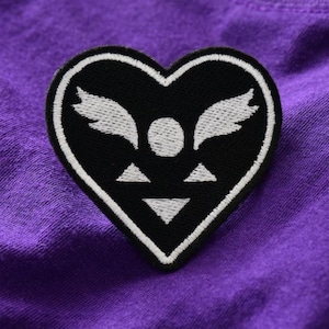 May include: Black heart-shaped patch with white embroidered details, including wings, a circle, and triangles. The patch is set against a textured purple fabric background.