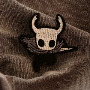 May include: Black and white embroidered patch of a knight character from the video game Hollow Knight. The knight is wearing a cloak and has a large, pointed helmet.