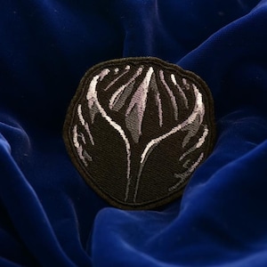 May include: A black patch with a stylized white and gray floral design. The patch is oval-shaped and has a black border. The background is a deep blue velvet fabric.