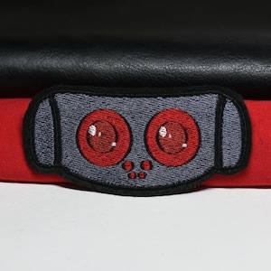 May include: A grey embroidered patch with two large red circles and five smaller grey circles. The patch is shaped like a robot head.