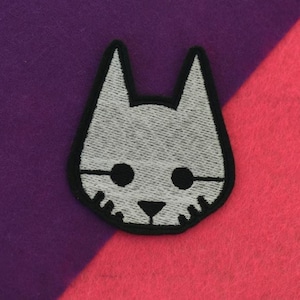 May include: A grey embroidered cat face patch with black eyes and whiskers. The patch is on a purple and pink background.