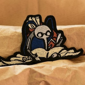 May include: Embroidered patch featuring a cartoon character wearing glasses and a backpack, surrounded by books. The character is white with black eyes and a blue and red backpack. The patch is black with white and beige details.