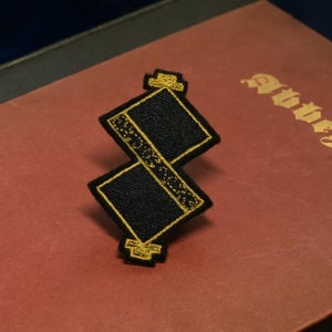 May include: A black and gold embroidered patch in the shape of a stylised 'S' with a gold border. The patch is on a red book with the word 'Abbeys' embossed in gold.