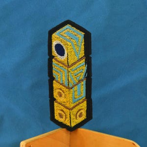May include: An embroidered patch featuring a yellow and blue totem pole with a black outline. The totem pole has a circular design at the top and is made of stacked blocks.