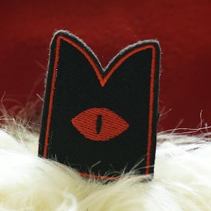 May include: Black fabric patch with a red eye design. The patch has a red outline and is sewn with a black thread.