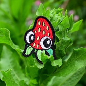 May include: An embroidered patch of a cartoon strawberry with googly eyes. The strawberry is red and green and is on a green leafy background.