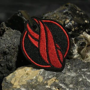 May include: A black and red embroidered patch with a stylised flame design. The patch is circular and has a black border.